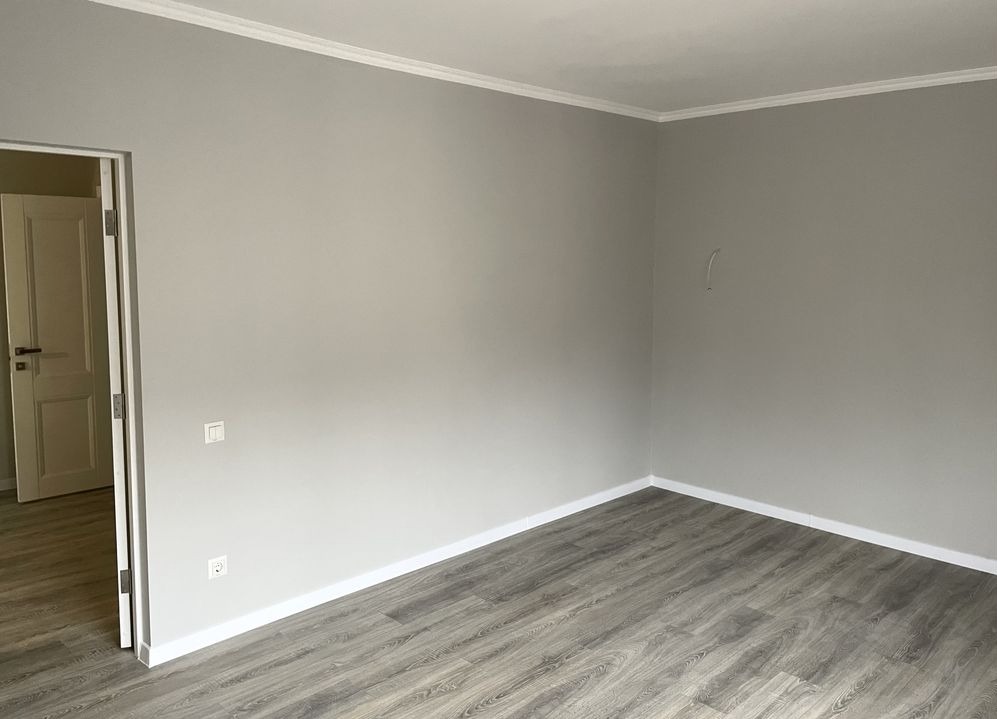 Pomalowane na szaro wnętrze pokoju z szarymi panelami podłogowymi i białymi listwami przypodłogowymi oraz sufitowymi, widoczne gniazdko elektryczne i włącznik światła.