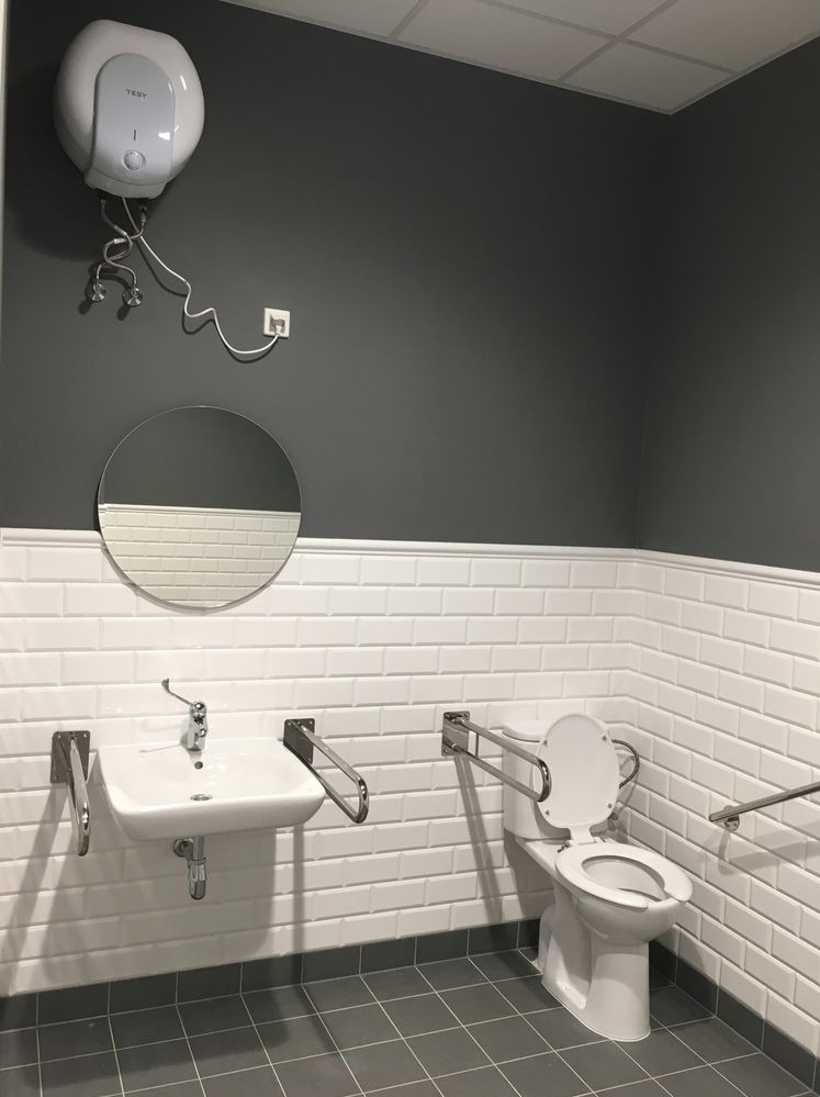 Wykończona łazienka z szarymi płytkami podłogowymi, białymi płytkami ściennymi typu metro, umywalką, toaletą z uchwytami dla niepełnosprawnych i okrągłym lustrem na szarej ścianie, nad umywalką...