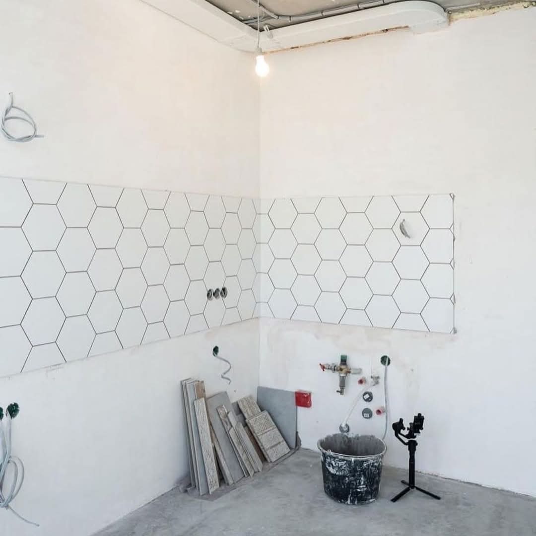 Białe heksagonalne płytki częściowo ułożone na ścianach w rogu jasnego pomieszczenia w trakcie remontu, widoczne instalacje, wiadro i poziomica na trójnogu.