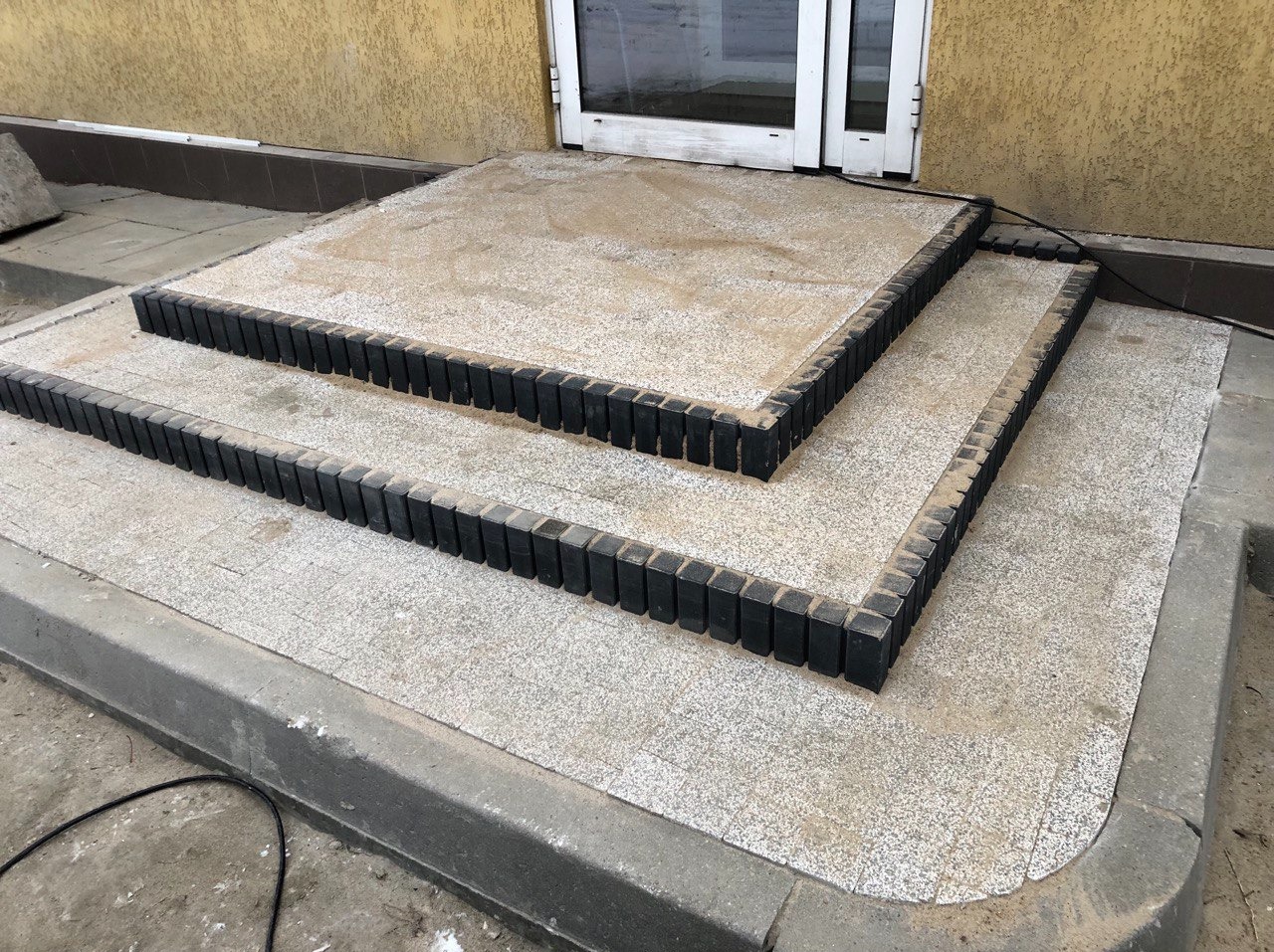 Wykończone schody wejściowe z jasnoszarej terakoty obramowane czarnymi kostkami, prowadzące do drzwi wejściowych.