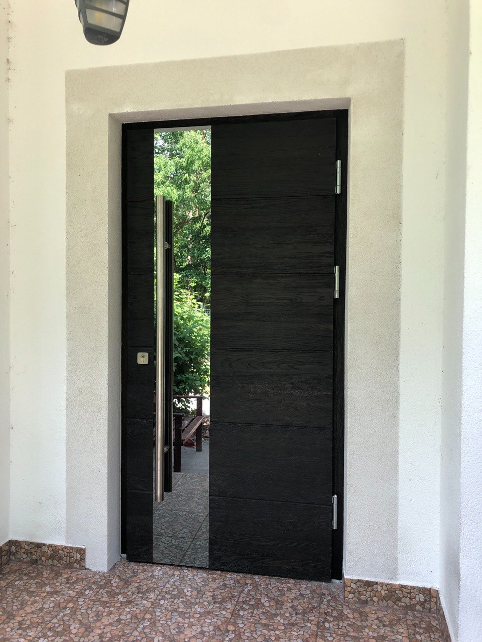 Częściowo otwarte, nowoczesne, czarne drzwi wejściowe z pionowym, lustrzanym panelem, osadzone w białym, otynkowanym portalu, widok na zieleń ogrodu.