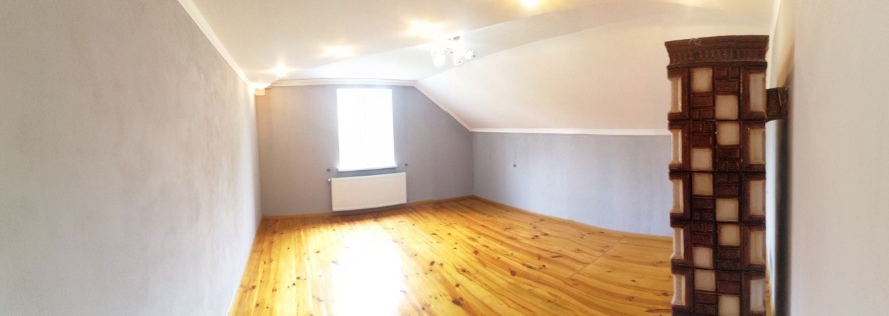 Pokój na poddaszu po remoncie: szare ściany, drewniana podłoga, biały grzejnik pod oknem i piec kaflowy z brązowo-beżowymi kaflami.