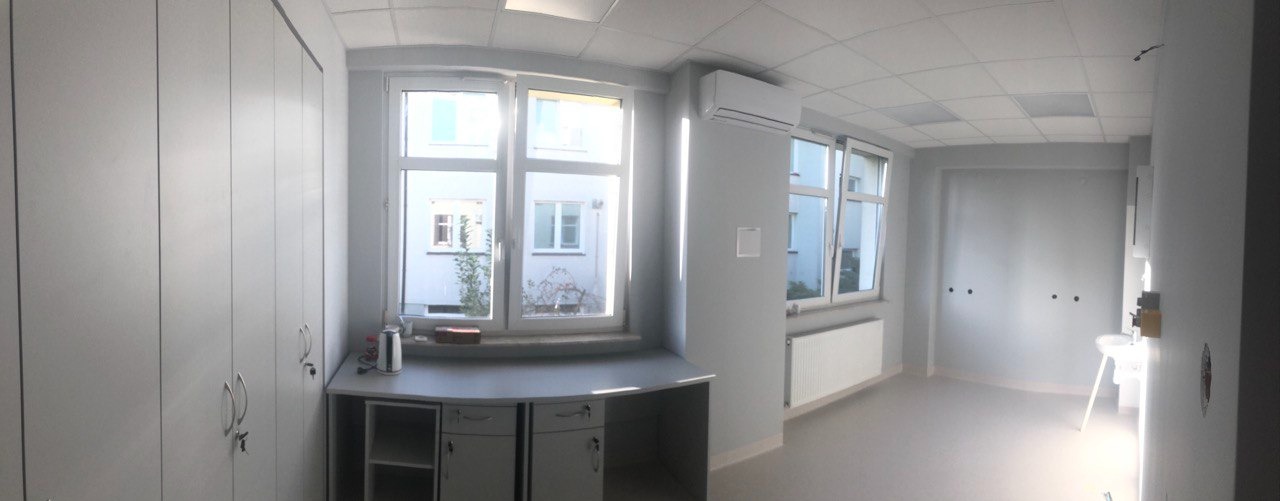 Wyremontowane, jasne wnętrze biurowe z szarymi ścianami, jasną podłogą, białymi szafami w zabudowie, biurkiem pod oknem i klimatyzacją.