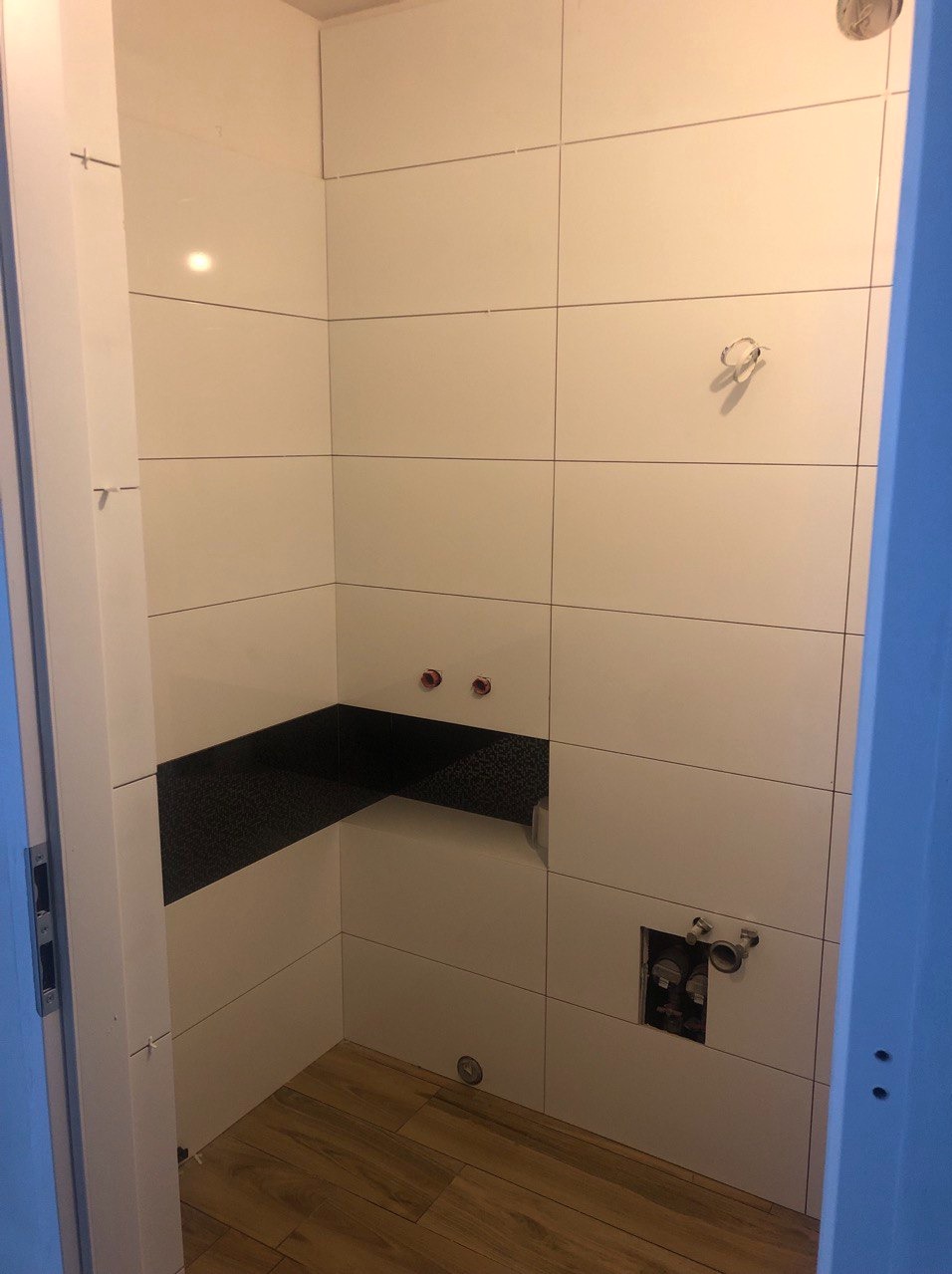 Wnętrze łazienki w trakcie wykańczania: białe płytki na ścianach z czarnym pasem dekoracyjnym, drewnopodobne panele na podłodze, widoczne wyprowadzenia instalacji wodnej i kanalizacyjnej.