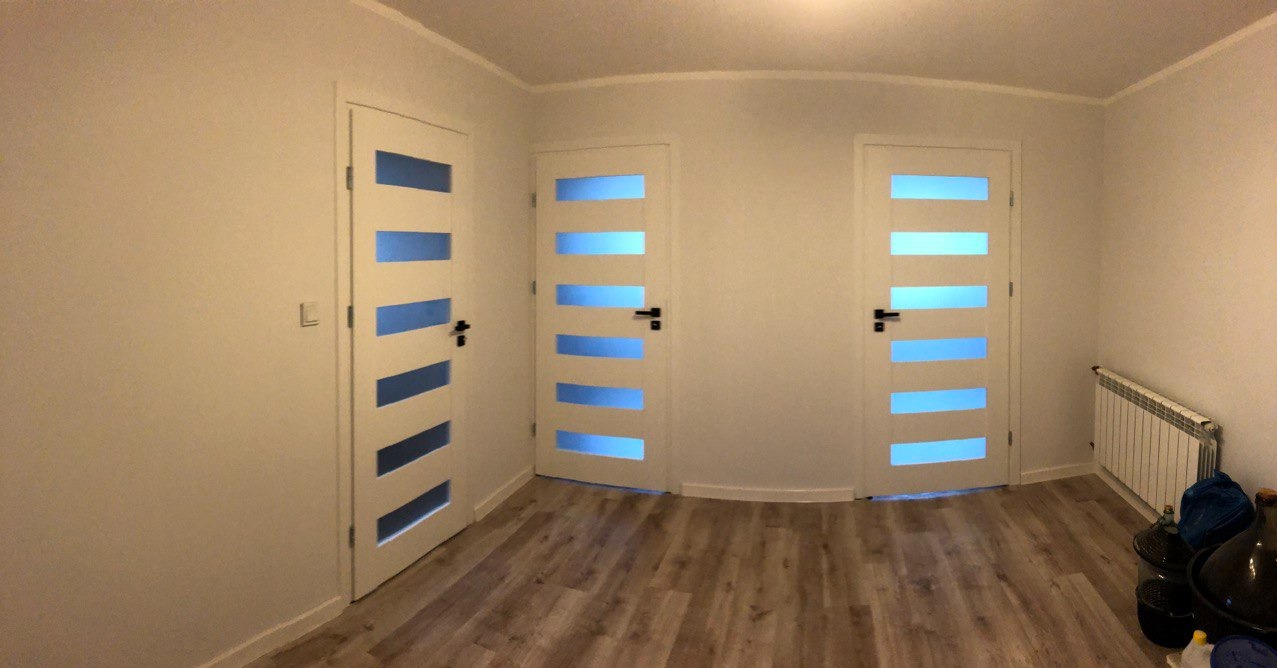 Wnętrze po remoncie: trzy białe drzwi z poziomymi, niebieskimi przeszkleniami, brązowe panele podłogowe, białe ściany i grzejnik.