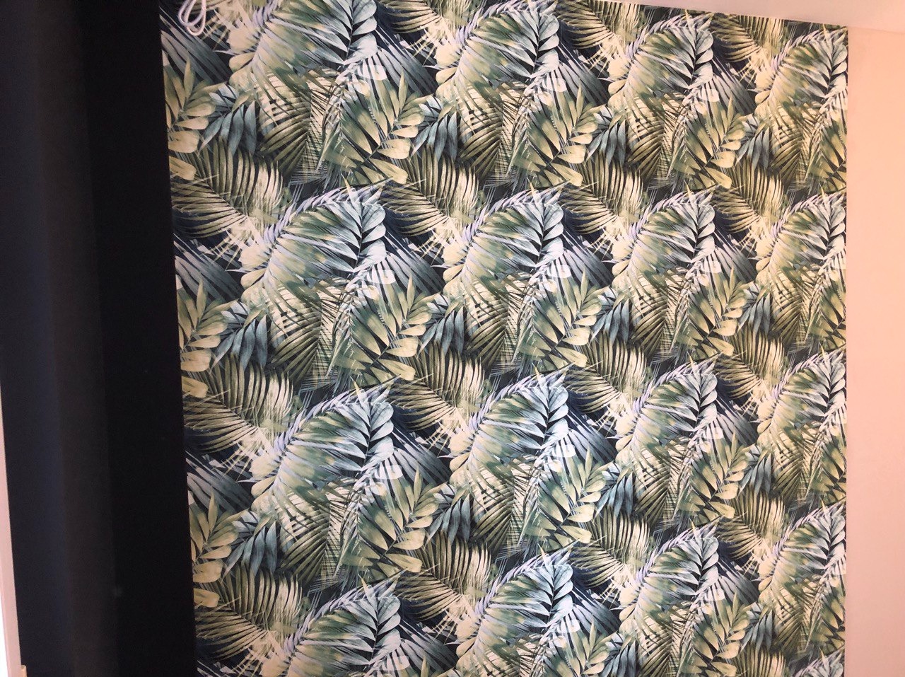 Ściana w Piastowie pokryta tapetą z motywem liści palmowych w odcieniach zieleni i błękitu, sąsiadująca z czarną zasłoną i fragmentem jasnoróżowej ściany.