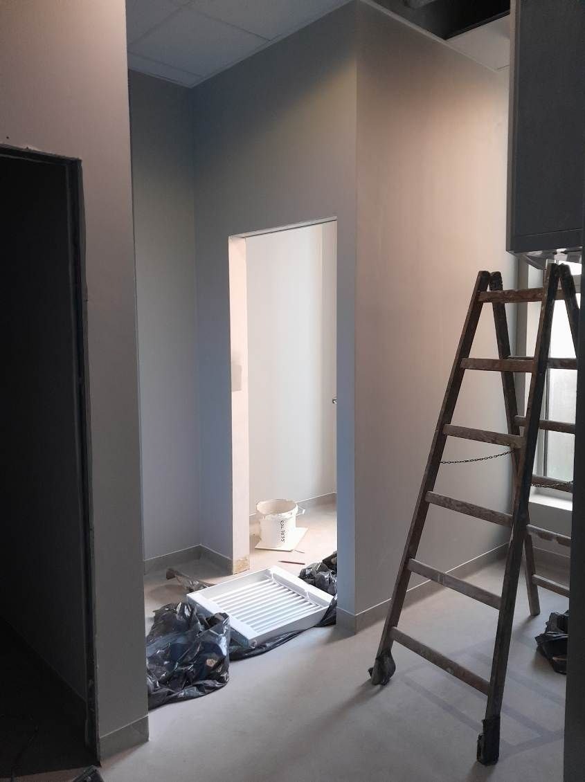 Świeżo pomalowane na szaro ściany w pomieszczeniu, widoczna drewniana drabina, kuweta malarska i wiadro z farbą, pomieszczenie w trakcie prac wykończeniowych.