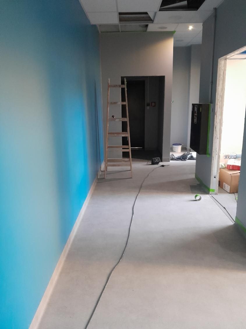 Wnętrze w trakcie prac wykończeniowych: ściana pomalowana na niebiesko, surowy beton na podłodze, drabina oparta o ścianę, widoczne ślady taśmy malarskiej, kabel na podłodze.