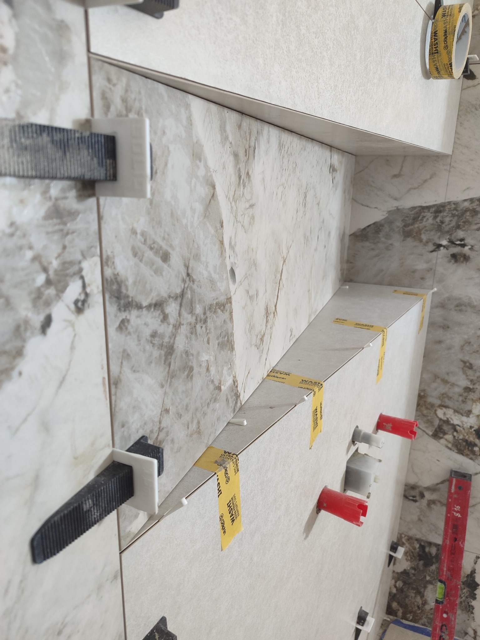 Układanie płytek imitujących marmur na ścianie, z użyciem klipsów poziomujących i taśmy maskującej. Widoczne otwory na armaturę i poziomica.