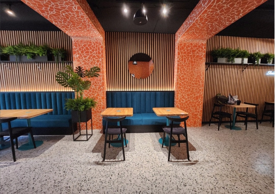 Wnętrze lokalu z mozaiką na filarach, drewnianymi panelami ściennymi, turkusowymi kanapami i stolikami. Podłoga z lastryko. Rośliny doniczkowe dodają świeżości.