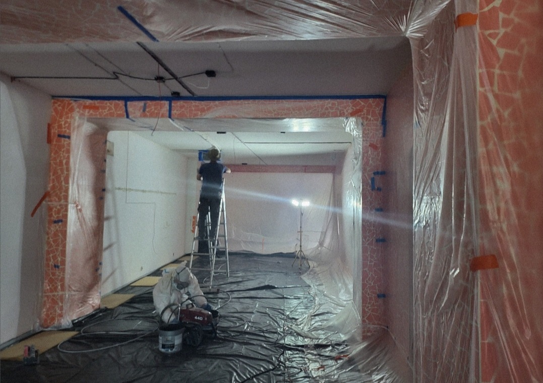 Prace przygotowawcze do remontu w Piastowie: zabezpieczone folią ściany z mozaiką, pracownik na drabinie i osoba w kombinezonie przy maszynie do natrysku.