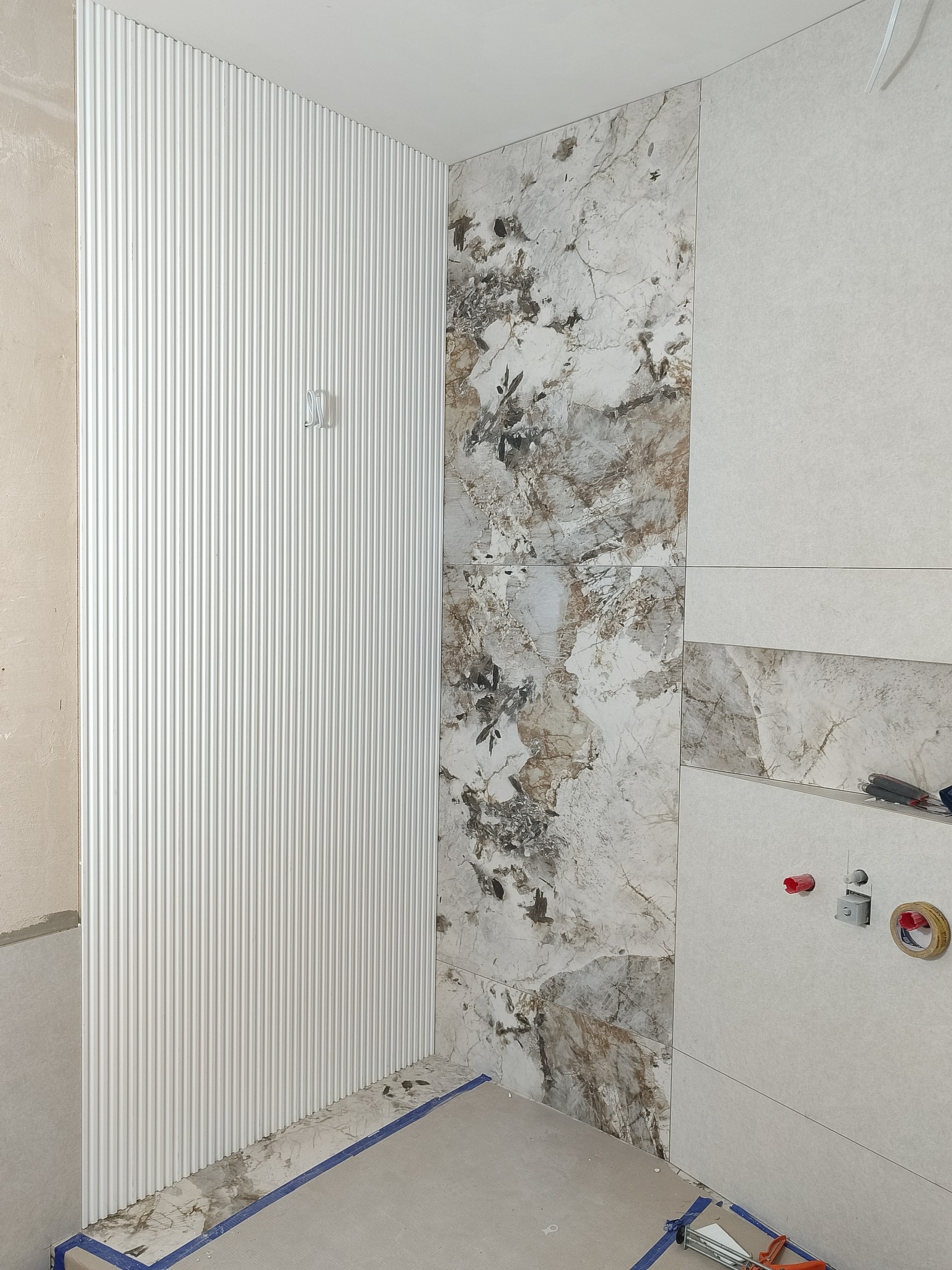Ściana z dekoracyjnymi panelami pionowymi i fragmentem marmurowych płytek w łazience w trakcie wykończenia. Widoczne narzędzia i taśma malarska.