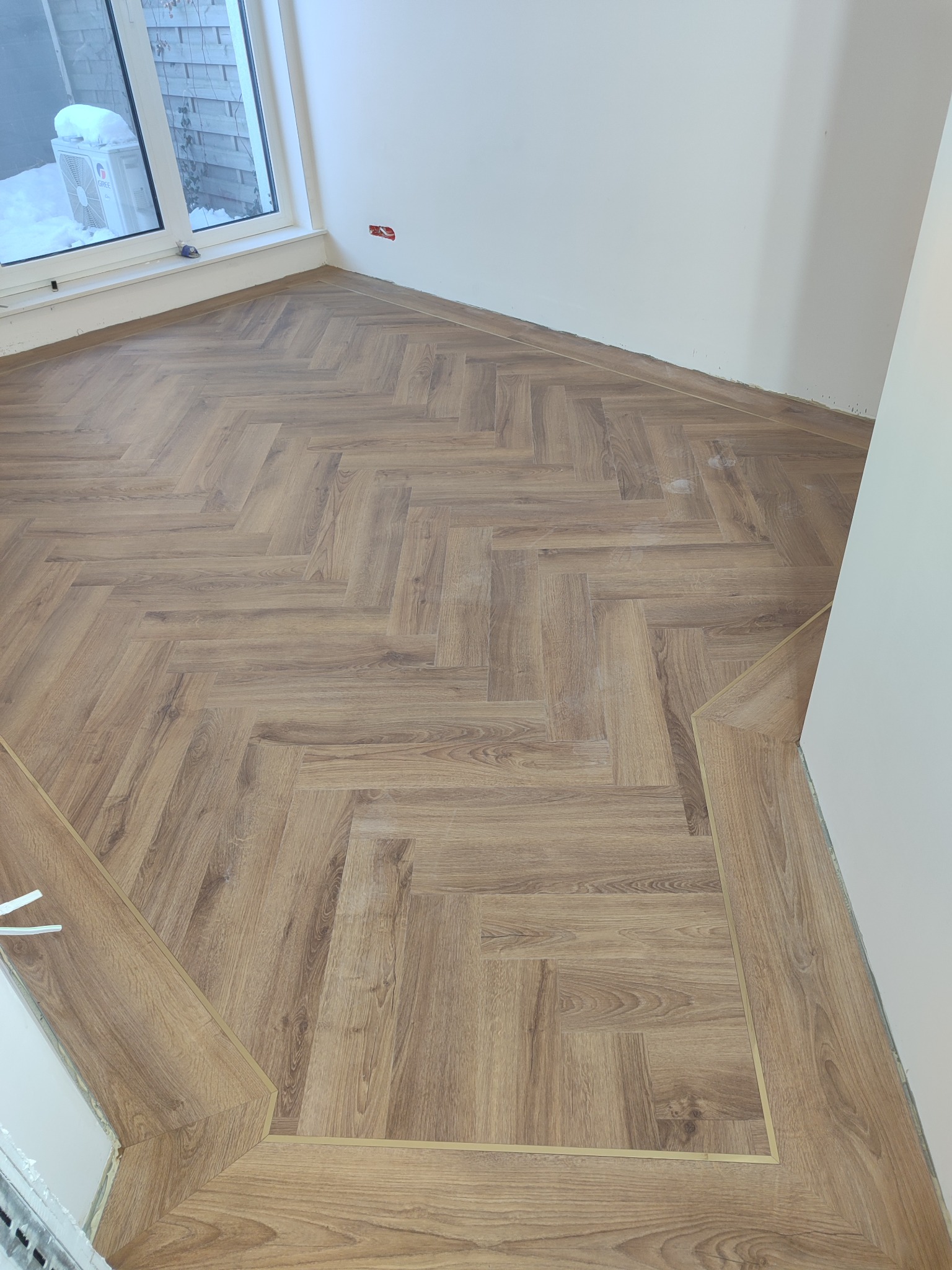 Ukończona podłoga z paneli ułożonych w jodełkę, z widocznymi listwami przypodłogowymi i fragmentem okna z widokiem na ośnieżony ogród.