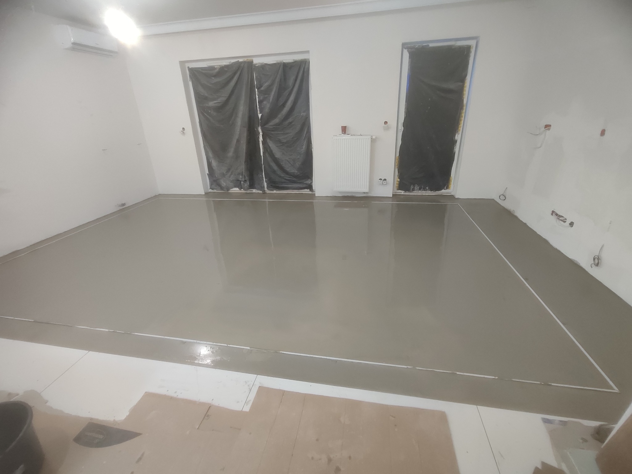 Wylany, świeży podkład podłogowy w jasnym pomieszczeniu z zabezpieczonymi folią oknami i drzwiami. Widoczne fragmenty starej podłogi i wiadro.
