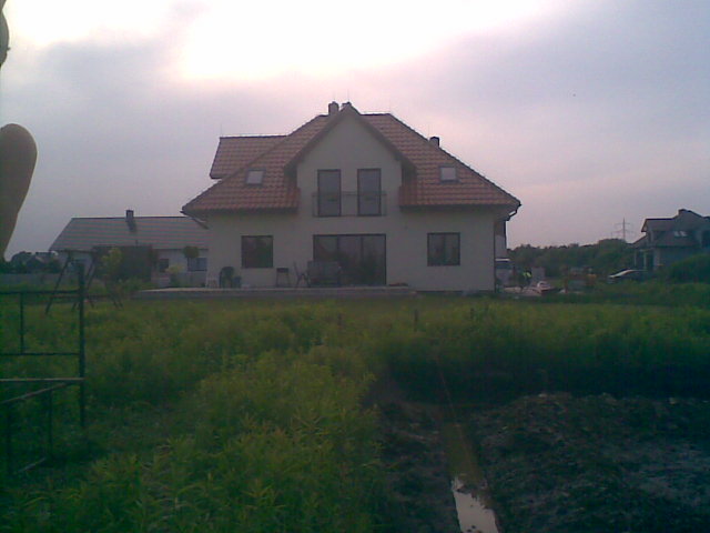 Niedokończony teren przed domem jednorodzinnym z czerwonym dachem, widoczne wykopane rowy i bujna roślinność.