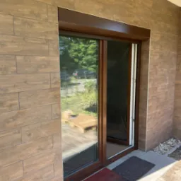 Brązowa rama okienna z moskitierą w nowoczesnym domu z okładziną imitującą drewno, widoczny fragment rolety zewnętrznej i oświetlenie zewnętrzne.