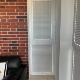 Białe plisy zamontowane w oknie balkonowym, widoczne na tle ceglanej ściany i fragmentu czarnej skórzanej sofy.