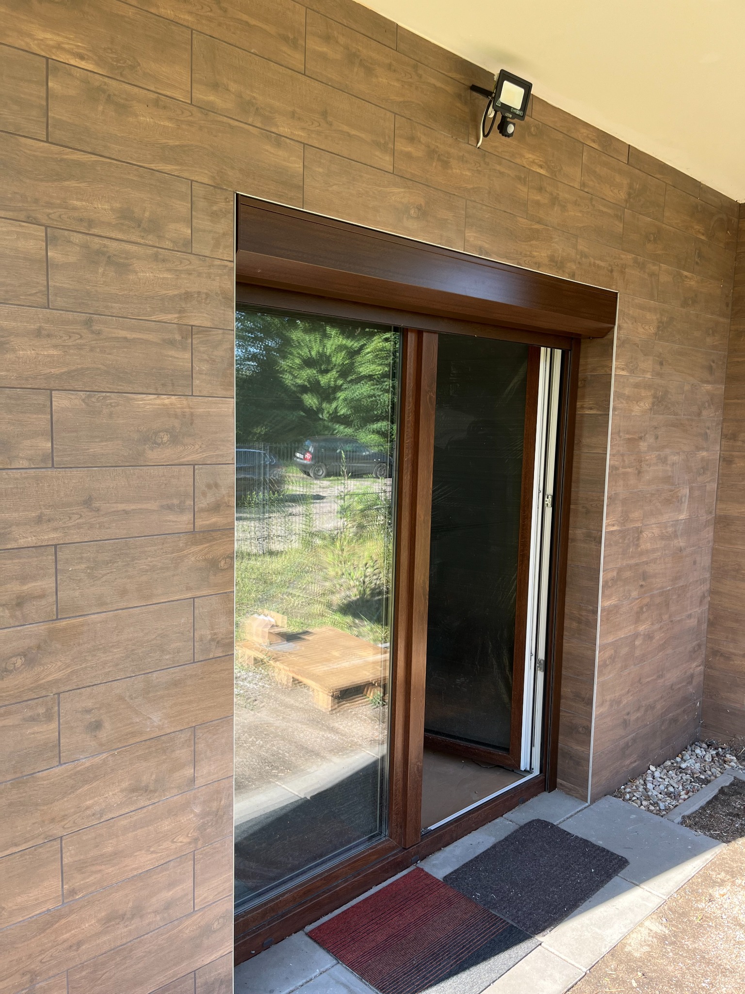 Zamknięta moskitiera ramkowa w brązowej ramie okiennej, widok z zewnątrz, brązowa elewacja imitująca drewno, czujnik ruchu nad oknem.