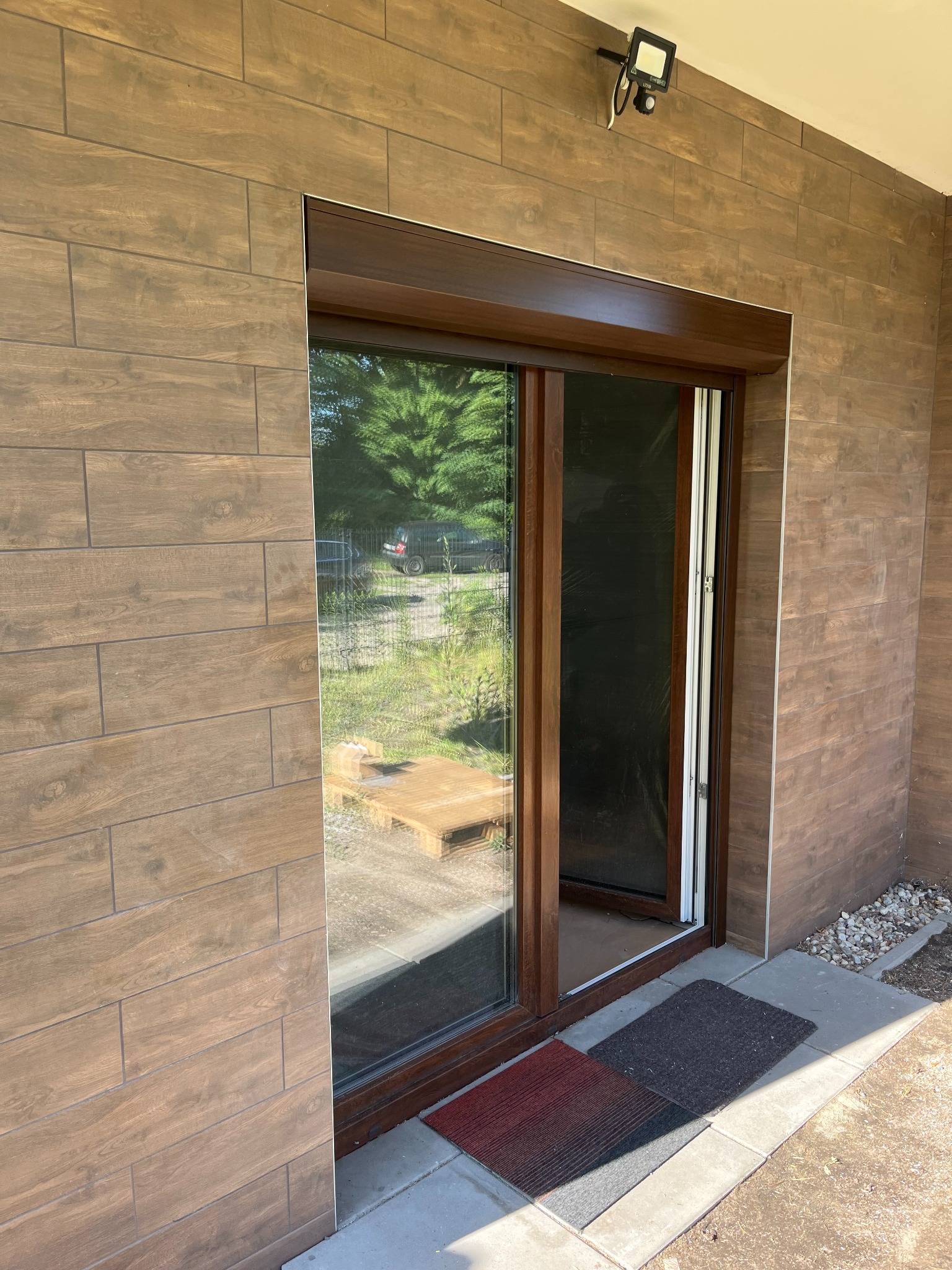 Brązowa rama okienna z moskitierą w nowoczesnym domu z okładziną imitującą drewno, widoczny fragment rolety zewnętrznej i oświetlenie zewnętrzne.