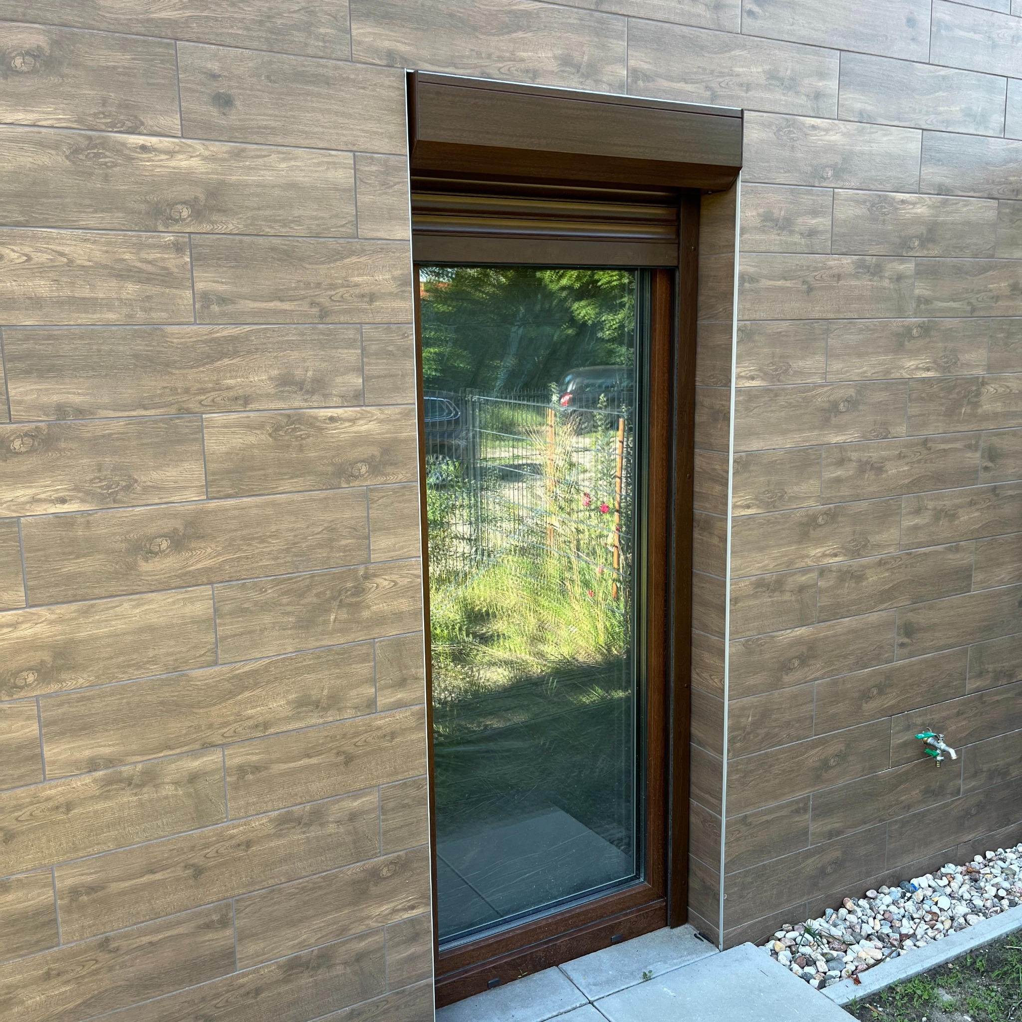 Brązowa roleta zewnętrzna częściowo opuszczona na oknie budynku z elewacją z płytek imitujących drewno, widok na ogród przez szybę.