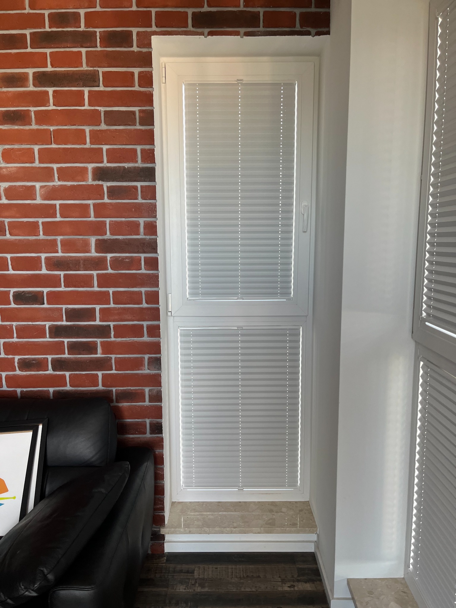 Białe plisy zamontowane w oknie balkonowym, widoczne na tle ceglanej ściany i fragmentu czarnej skórzanej sofy.