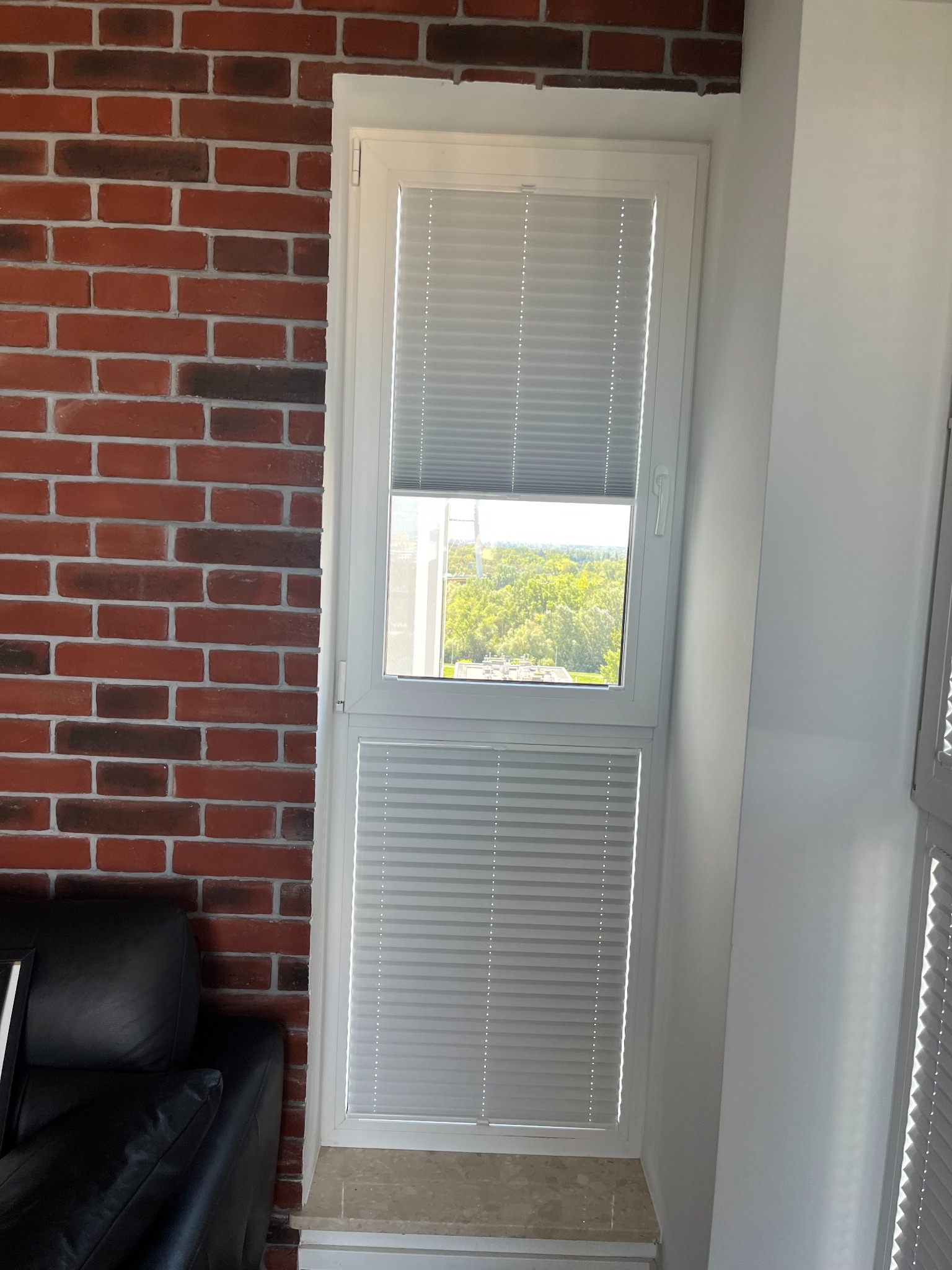 Białe okno z plisami podzielone na dwie części, dolna zasłonięta, górna odsłonięta z widokiem na zieleń, obok ściana z czerwonej cegły i fragment czarnej sofy.