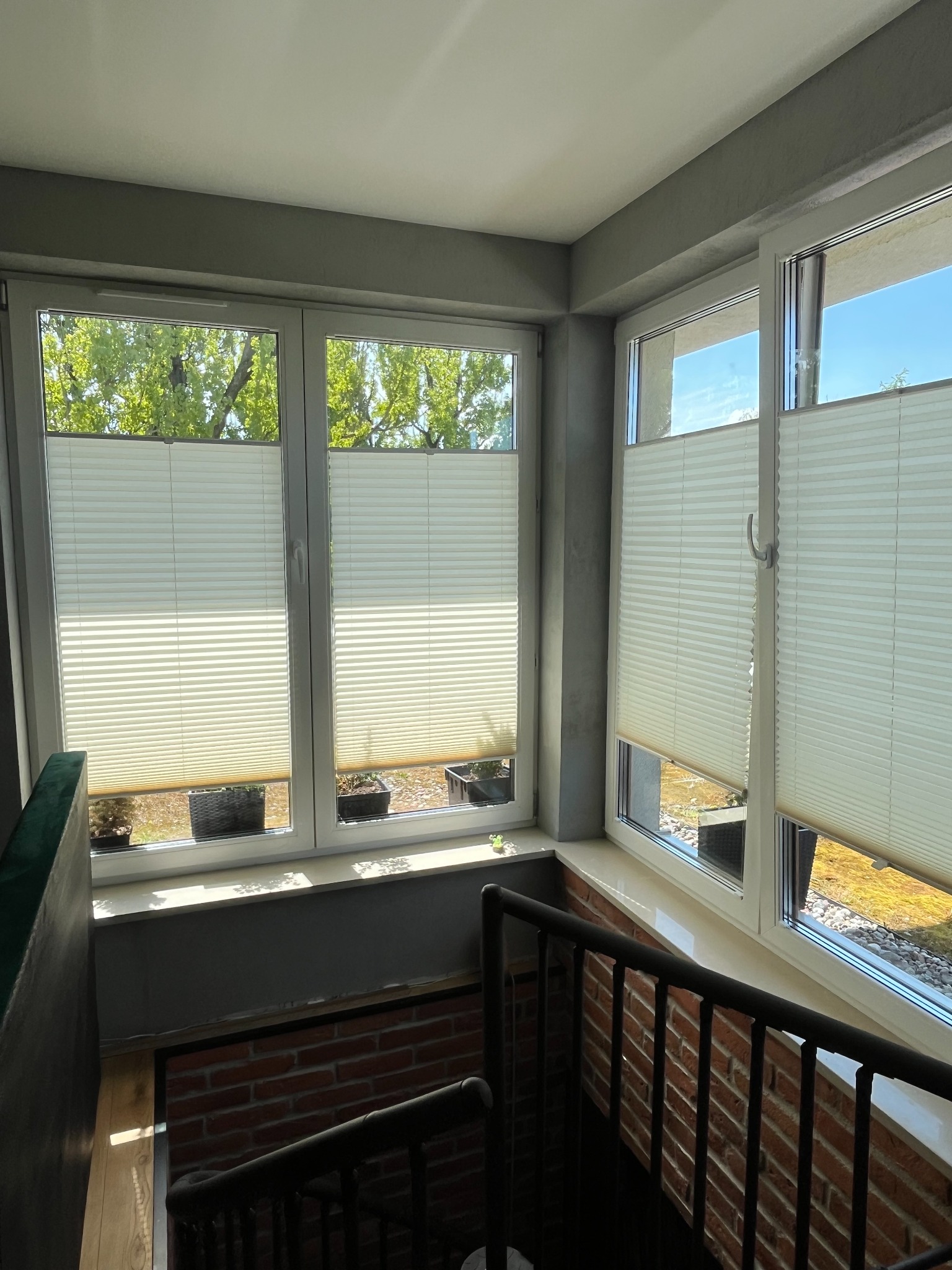 Jasne plisy okienne zamontowane w oknach na klatce schodowej w Warszawie, widok z dołu na częściowo zasłonięte okna z widokiem na zieleń.