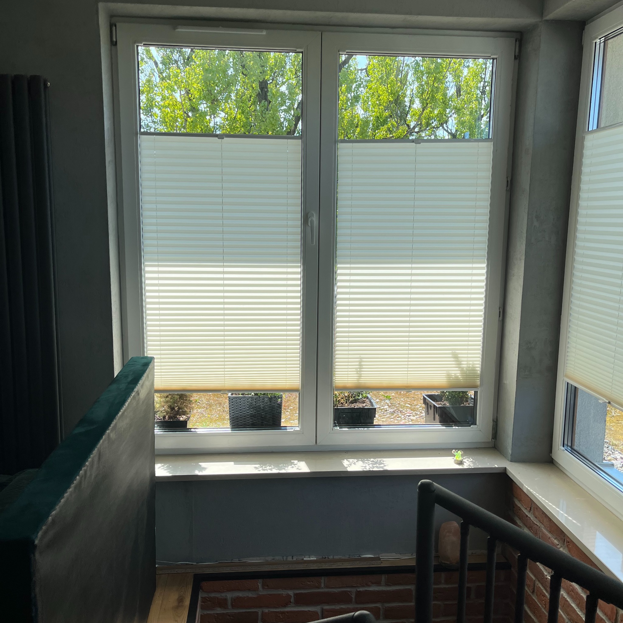 Jasne plisy okienne zamontowane w dwuskrzydłowym oknie PCV, widok z wnętrza na zieleń za oknem i doniczki z roślinami na parapecie, ujęcie z dołu schodów z ceglaną ścianą.