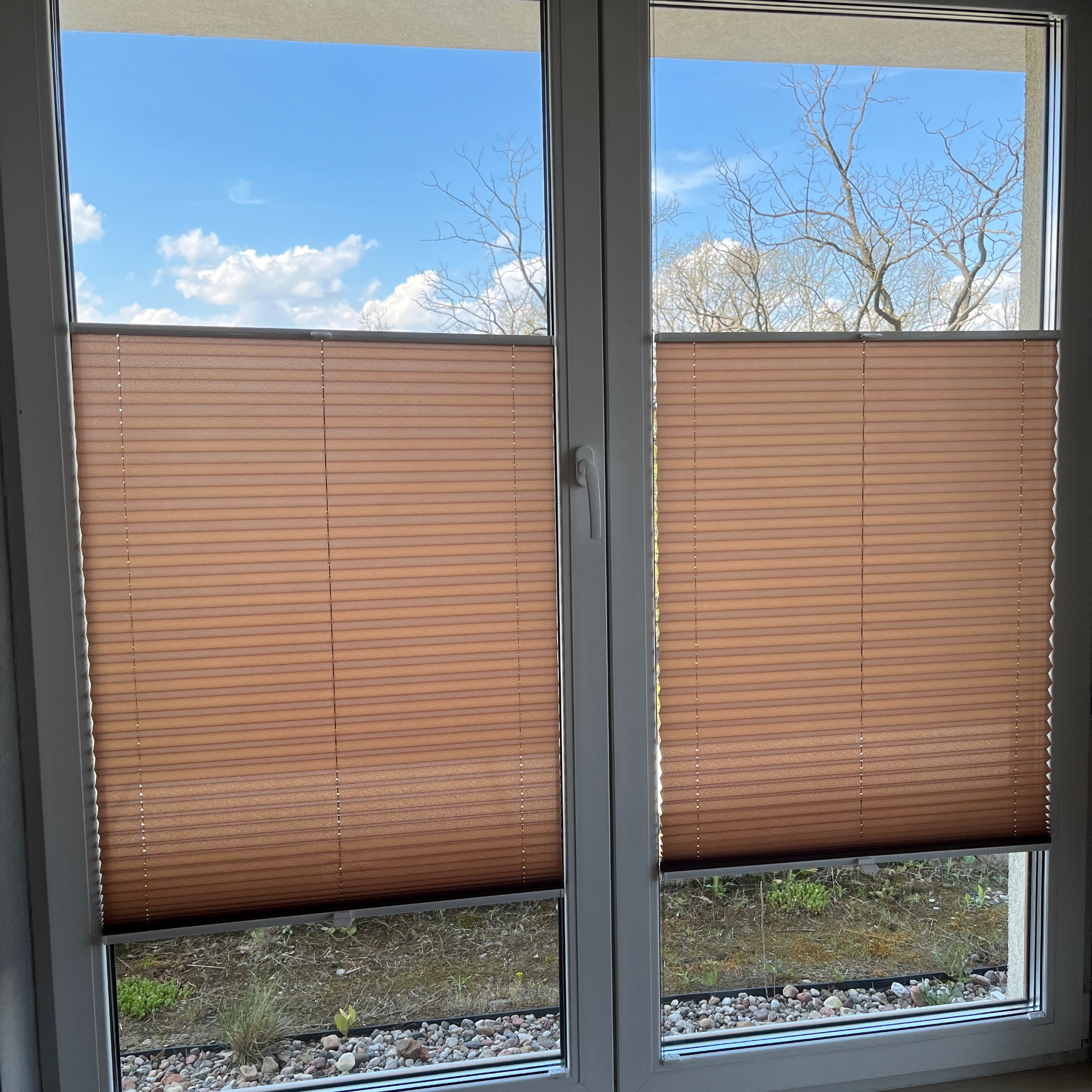 Brązowe plisy w oknie PCV z widokiem na ogródek skalny i błękitne niebo z chmurami.