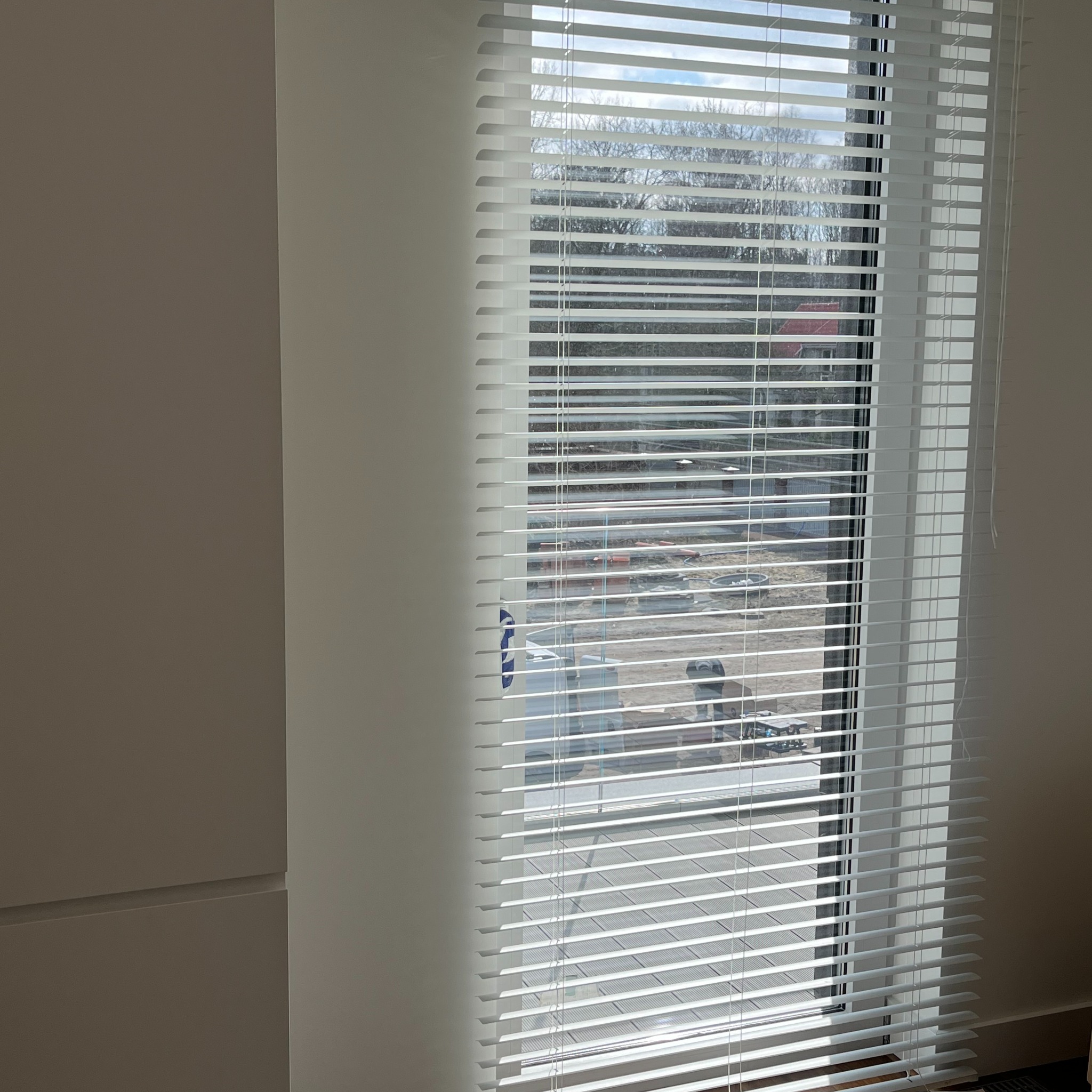 Białe, poziome żaluzje wewnętrzne zamontowane w oknie, częściowo zasłonięte, z widokiem na plac budowy na zewnątrz.