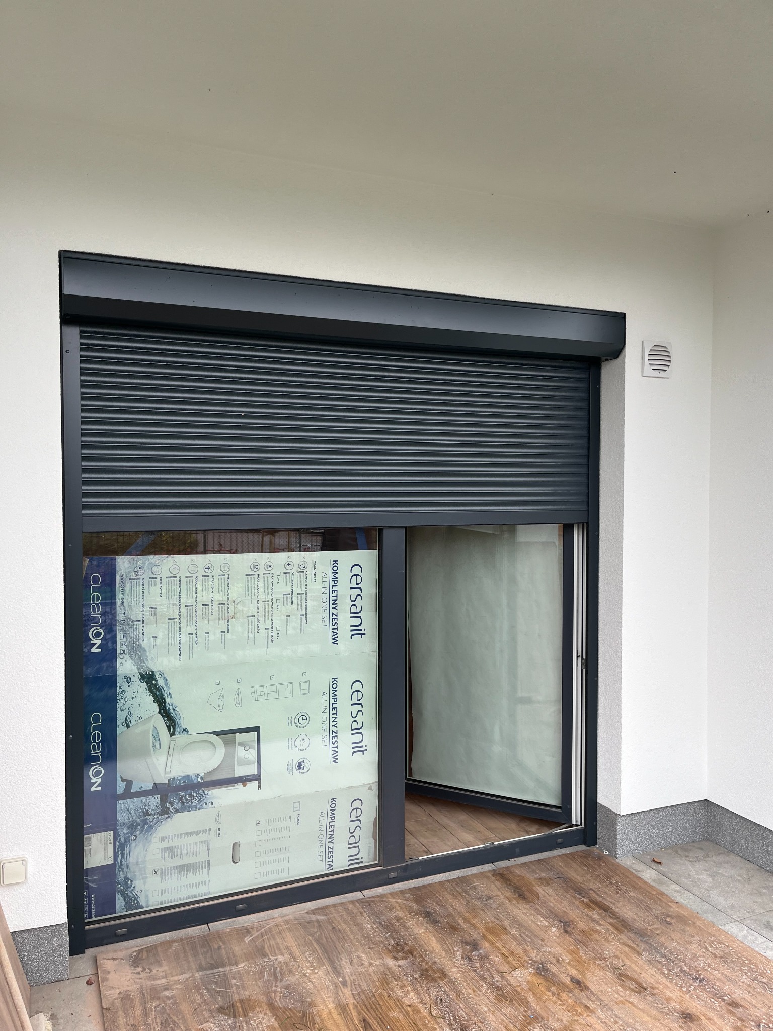 Zamknięta grafitowa roleta zewnętrzna na oknie tarasowym w Warszawie, widoczne kartony z wyposażeniem łazienki za szybą, drewniana podłoga tarasu.