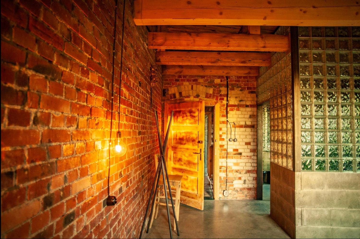 Wnętrze loftu z odsłoniętą cegłą, wiszącą żarówką na kablu, drewnianymi belkami stropowymi i ścianą z luksferów, ukazujące stylową instalację elektryczną.