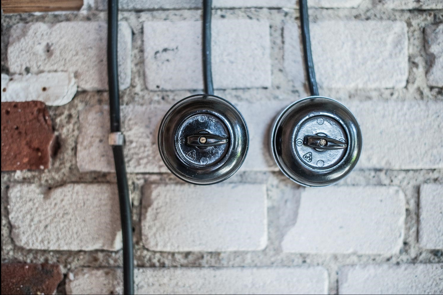 Dwa okrągłe, czarne włączniki światła z przełącznikami obrotowymi zamontowane na ścianie z białej cegły z widocznymi przewodami elektrycznymi.