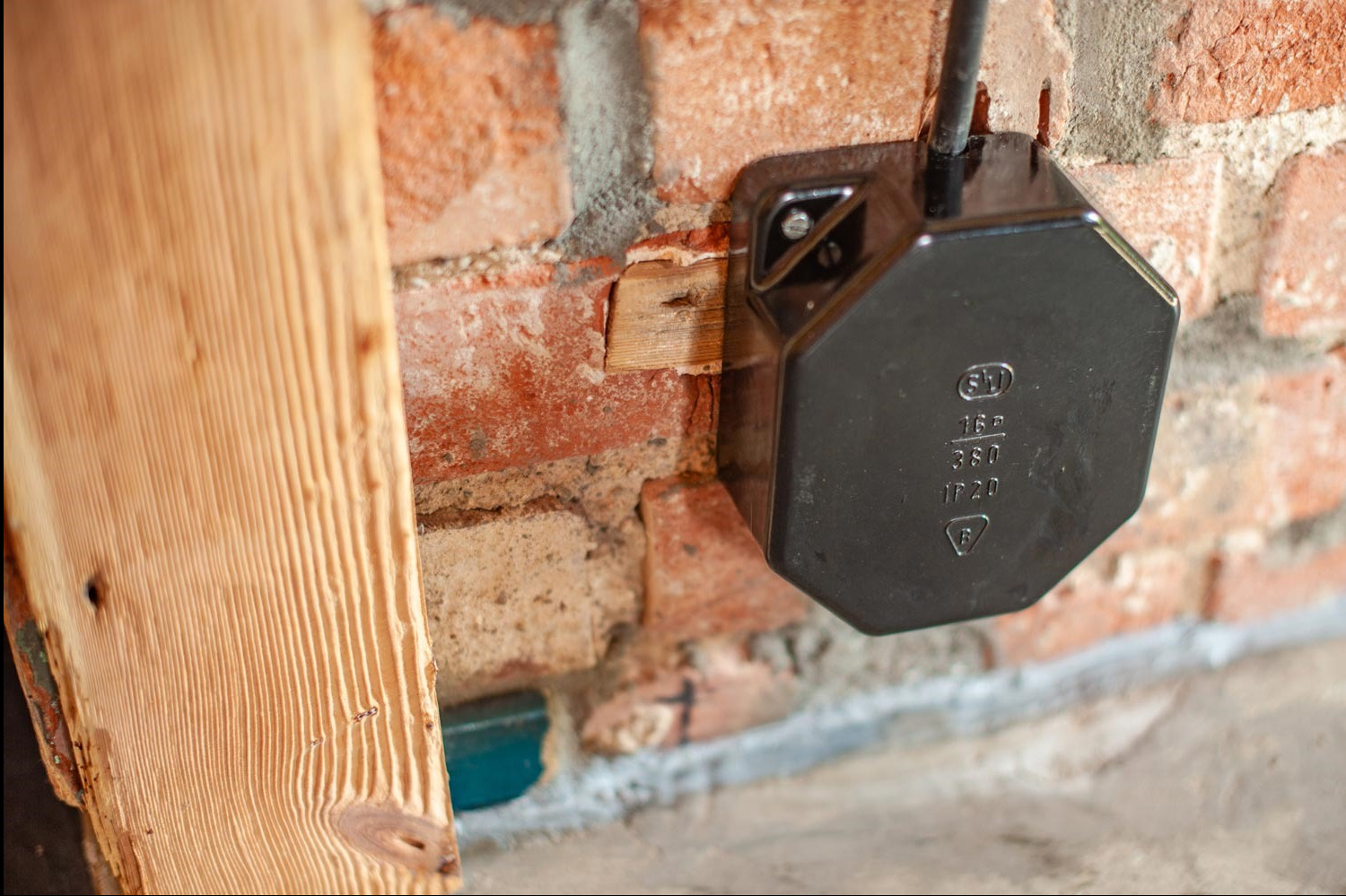 Czarna puszka elektryczna natynkowa zamontowana na ceglanej ścianie w Koninie, widoczne oznaczenia na puszce i fragment drewnianej listwy.