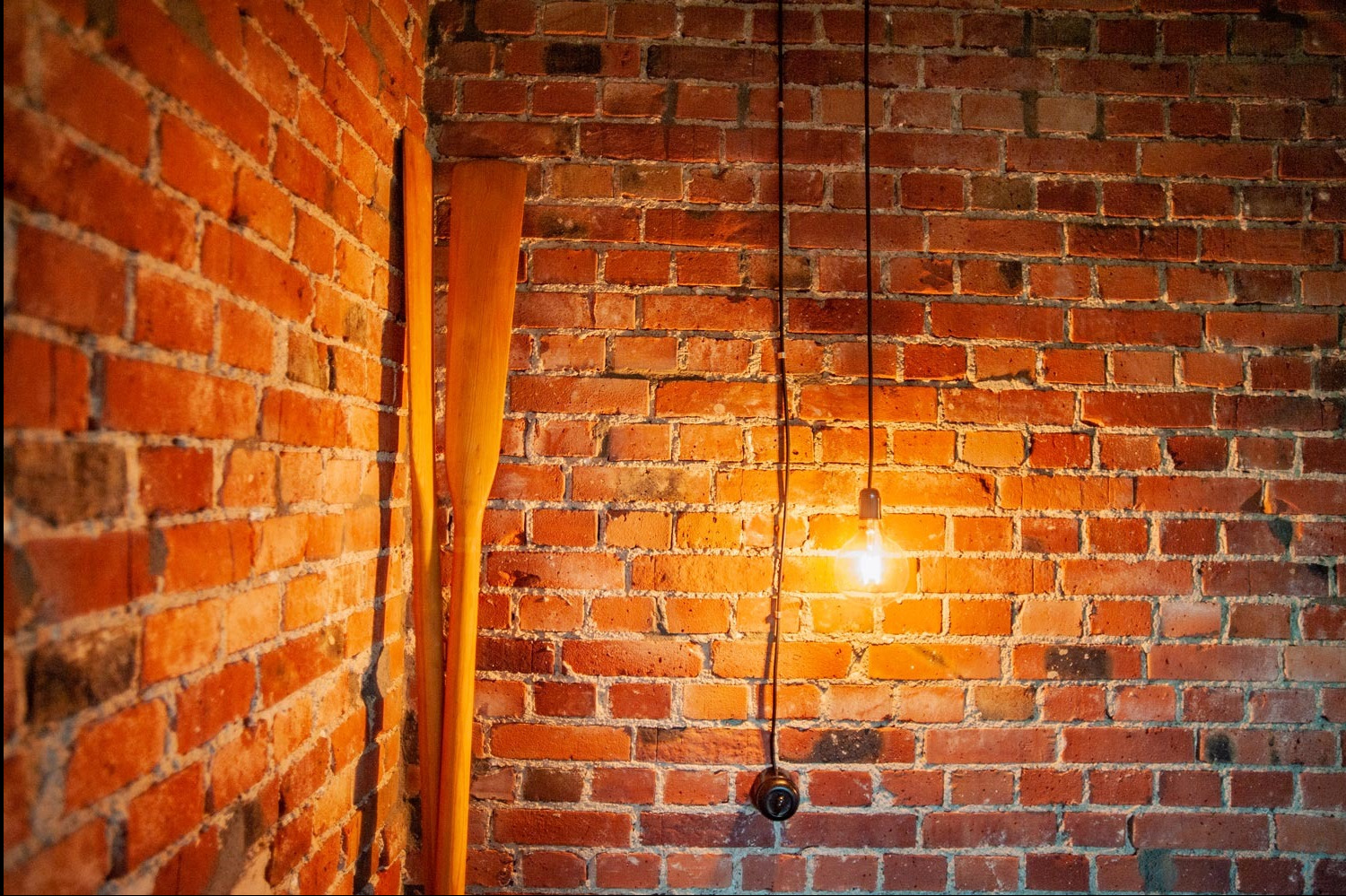 Żarówka Edisona zawieszona na kablu zwisającym wzdłuż ściany z czerwonej cegły, obok stoją dwie drewniane wiosła.