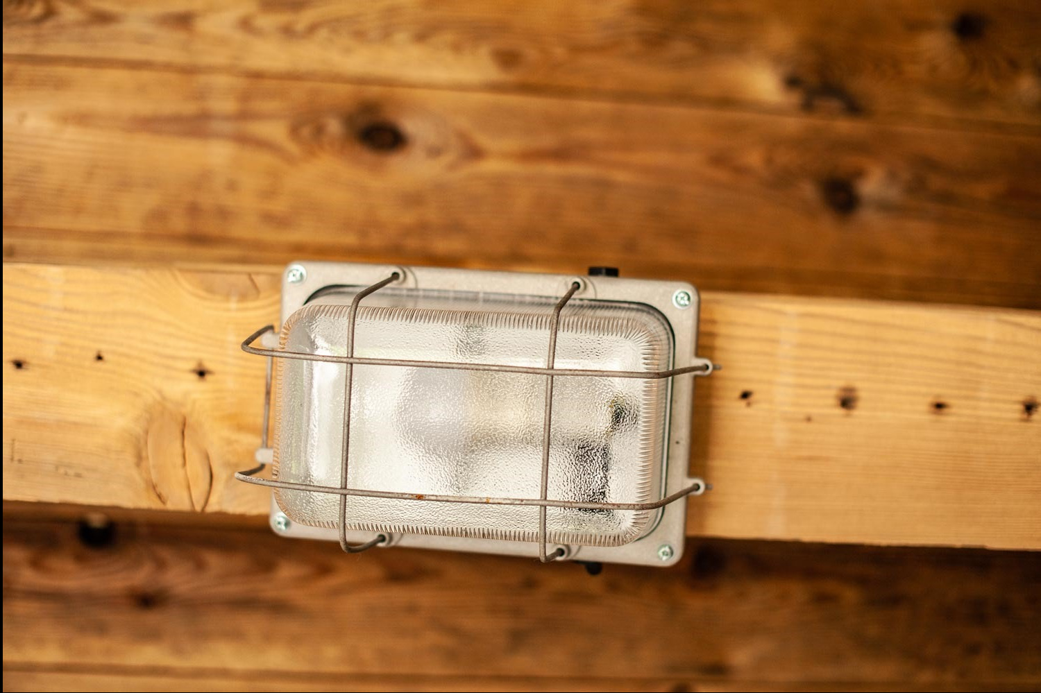 Zamknięta lampa przemysłowa z metalową kratką ochronną, zamontowana na drewnianej ścianie z widocznymi sękami i śladami po wkrętach.