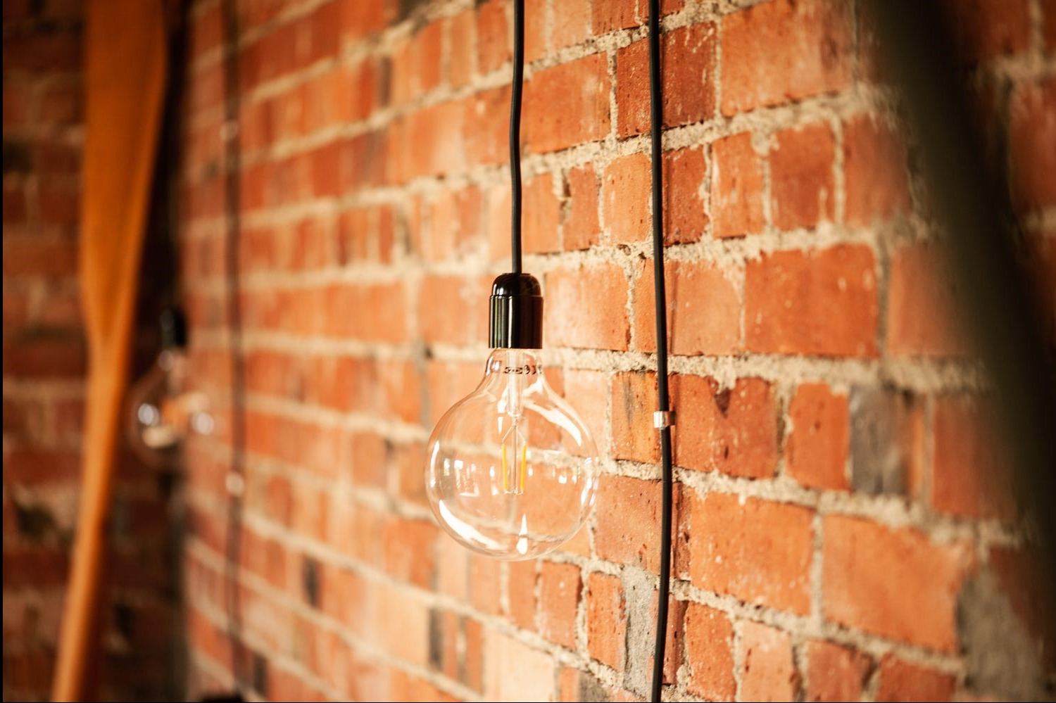 Żarówka dekoracyjna Edison z czarnym kablem zwisająca na tle ceglanej ściany w stylu loftowym, z widocznym drewnianym elementem w tle.