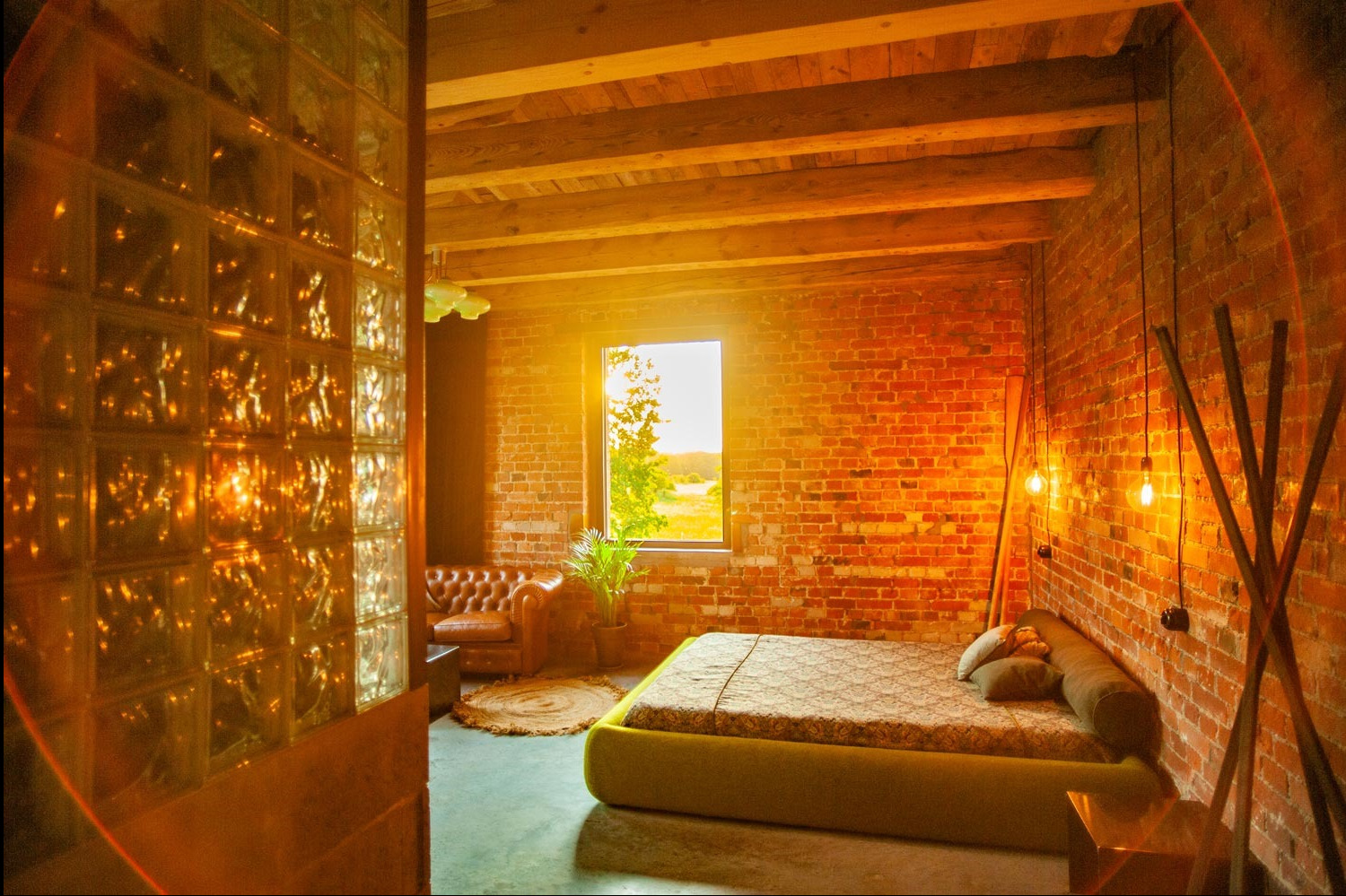 Loftowy apartament z ceglanymi ścianami, drewnianym stropem i szklaną ścianką działową, oświetlony ciepłym światłem słonecznym wpadającym przez okno. Widoczne wiszące żarówki na kablach...