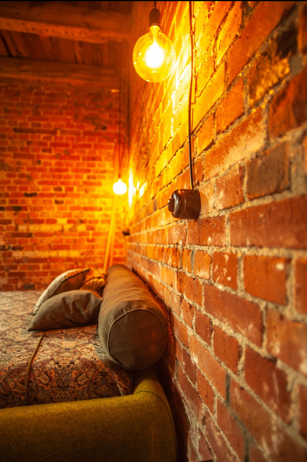 Loftowy apartament z ceglaną ścianą oświetloną żarówkami Edisona zwisającymi na kablach, widoczny włącznik elektryczny na ścianie i fragment łóżka z poduszkami.