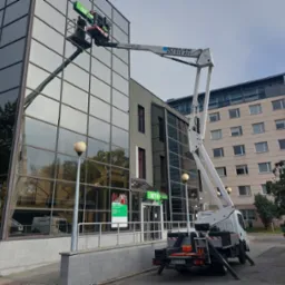 Pracownicy na podnośniku koszowym czyszczą szklaną fasadę budynku banku, widoczne narzędzia i akcesoria na platformie, odbicie drzew w szybach.