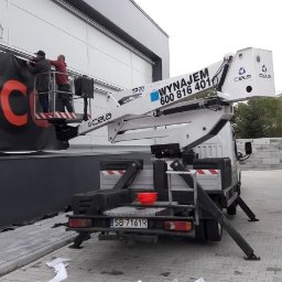 NAMI S.C - Montaż baneru reklamowego z czerwonym logo na elewacji budynku przy użyciu podnośnika koszowego Cela T220; widoczna drabina oparta o ścianę i elementy mocujące baner.