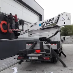 Montaż baneru reklamowego z czerwonym logo na elewacji budynku przy użyciu podnośnika koszowego Cela T220; widoczna drabina oparta o ścianę i elementy mocujące baner.