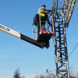 NAMI S.C - Pracownik w żółtej kamizelce ochronnej, w koszu podnośnika CELA TP20, pracuje na wysokości przy metalowej konstrukcji telekomunikacyjnej na tle błękitnego nieba.