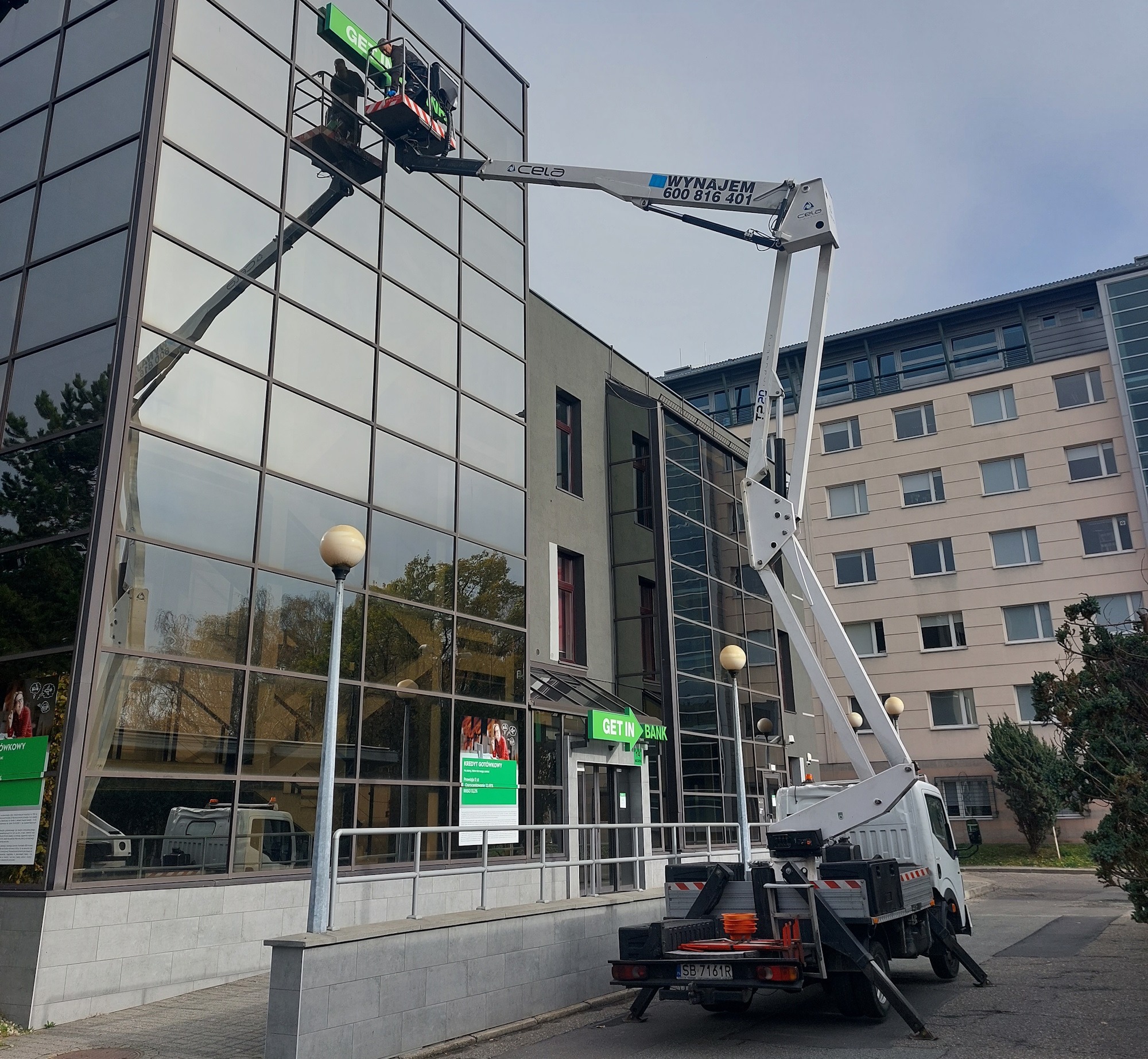 Pracownicy na podnośniku koszowym czyszczą szklaną fasadę budynku banku, widoczne narzędzia i akcesoria na platformie, odbicie drzew w szybach.