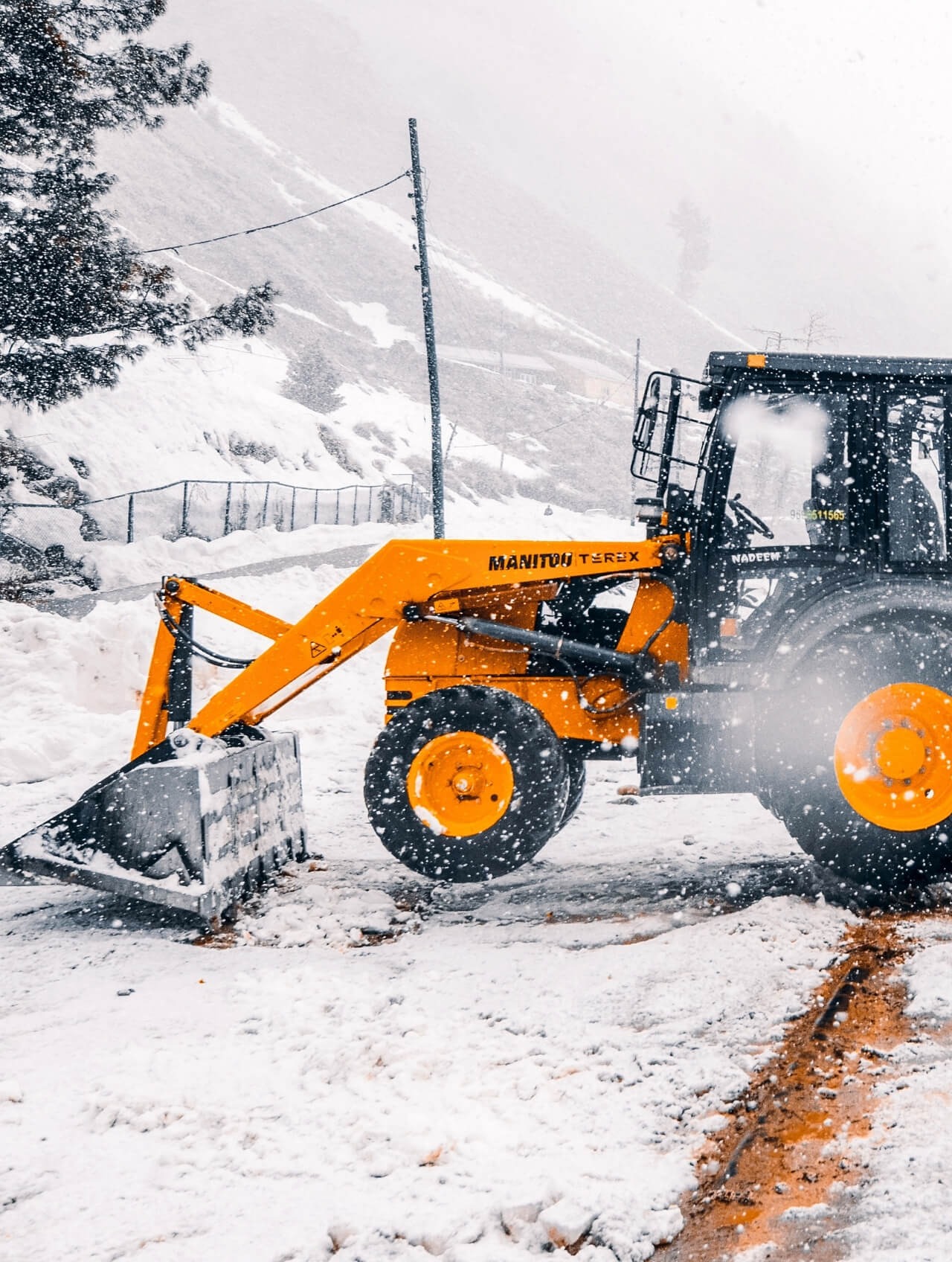 Żółto-czarna ładowarka Manitou Terex odśnieża teren w zimowej scenerii, z widocznymi płatkami śniegu i górą w tle.