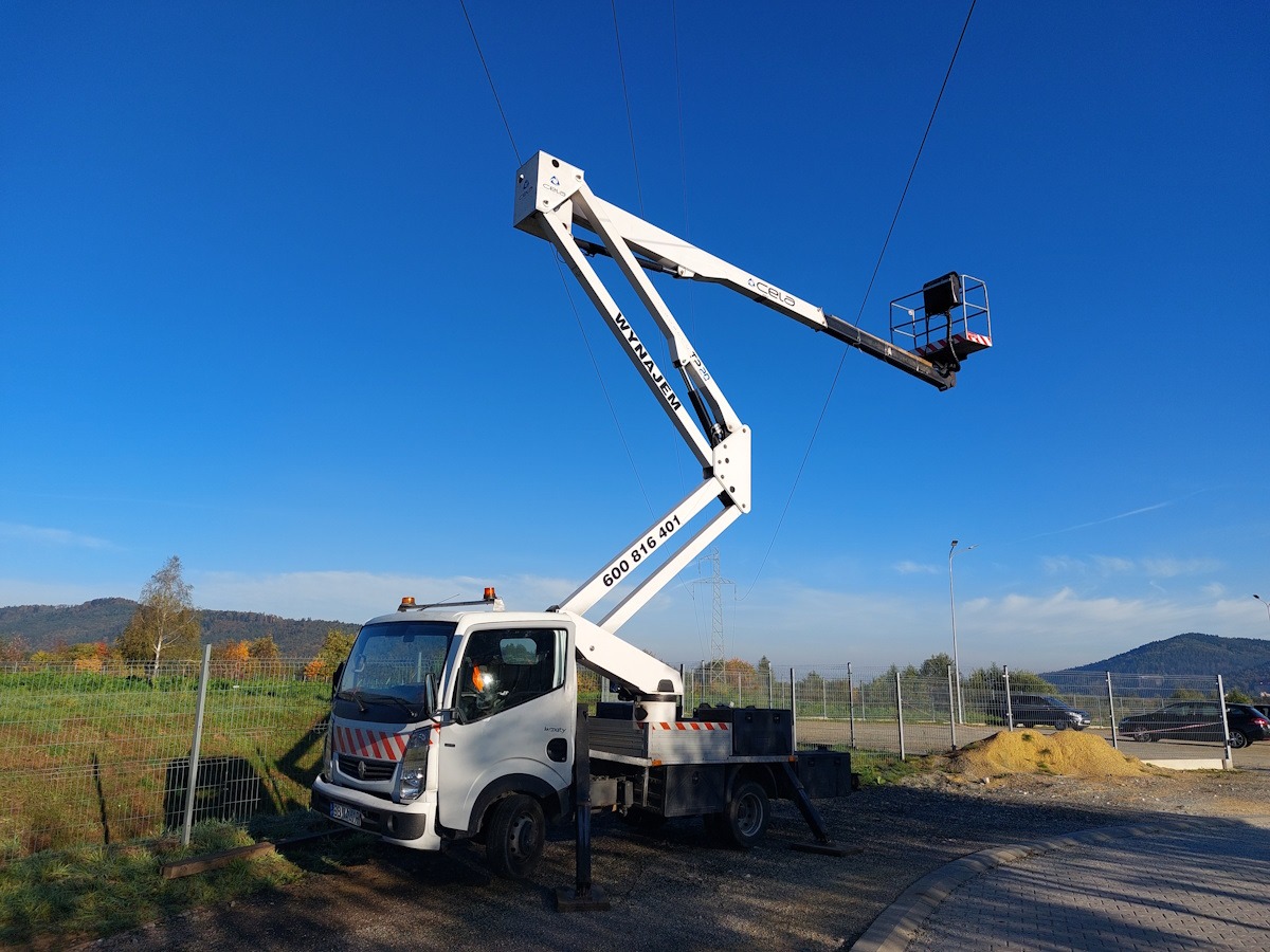 Biały samochód z wysięgnikiem teleskopowym na tle błękitnego nieba i zieleni, widoczne linie wysokiego napięcia
