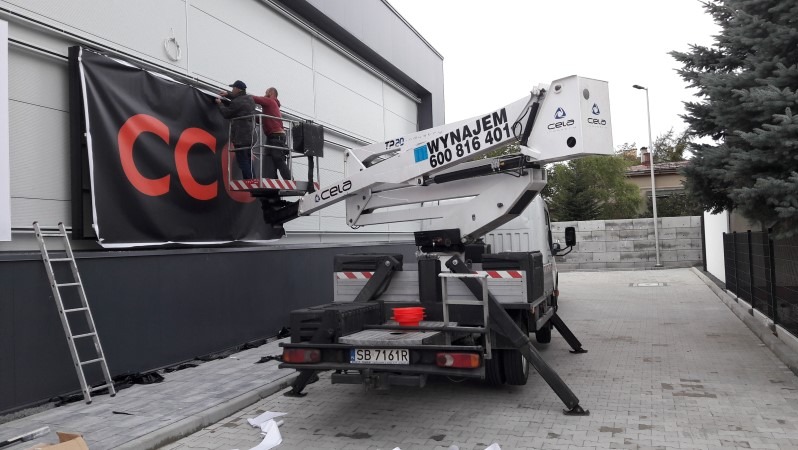 Montaż baneru reklamowego z czerwonym logo na elewacji budynku przy użyciu podnośnika koszowego Cela T220; widoczna drabina oparta o ścianę i elementy mocujące baner.