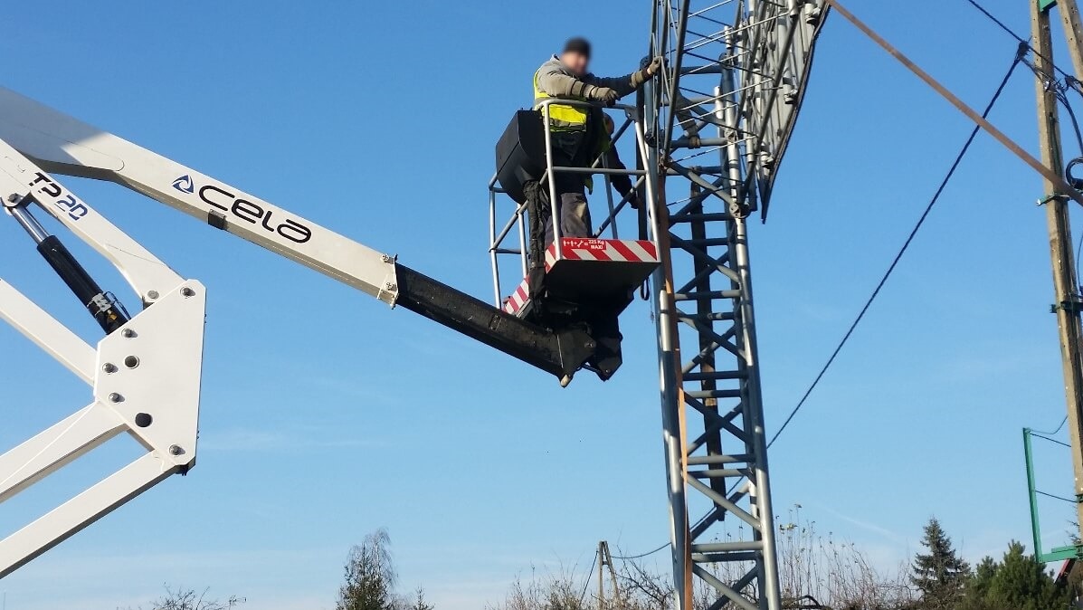 Pracownik w żółtej kamizelce ochronnej, w koszu podnośnika CELA TP20, pracuje na wysokości przy metalowej konstrukcji telekomunikacyjnej na tle błękitnego nieba.