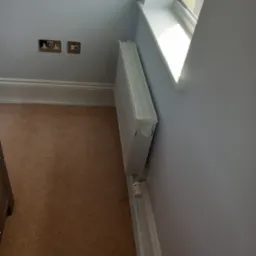 Świeżo pomalowane na szaro wnętrze z grzejnikiem zabezpieczonym taśmą malarską, gładkie ściany, brązowa podłoga i okno z białym parapetem.