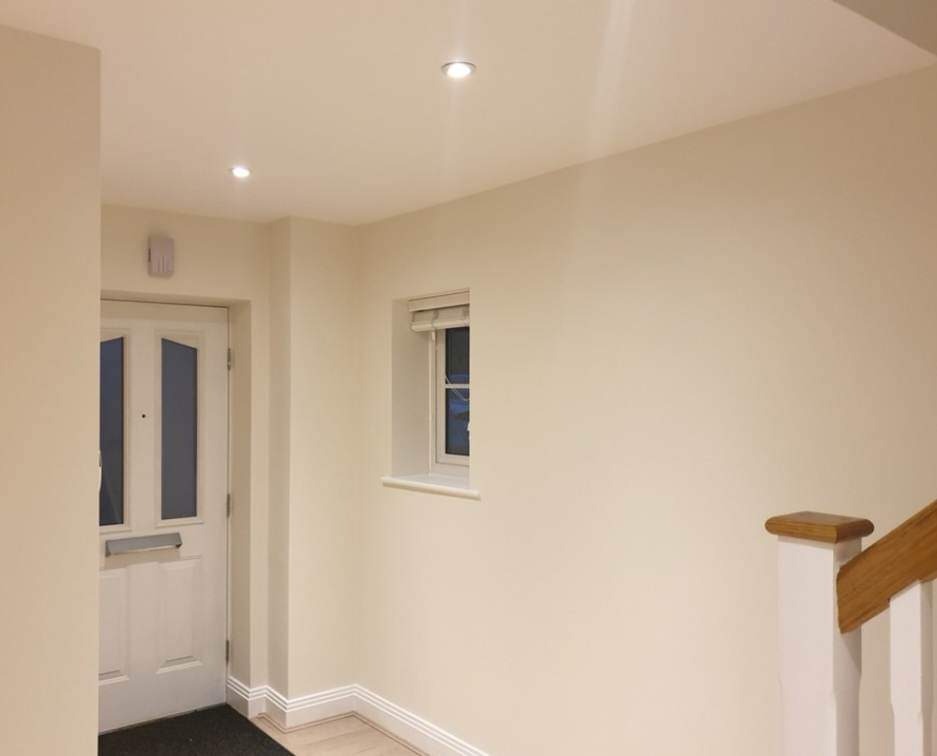 Jasne wnętrze po remoncie: świeżo pomalowane ściany w odcieniu kremowym, widoczny fragment klatki schodowej z drewnianą poręczą, białe drzwi wejściowe z przeszkleniami i małe okno z roletą.